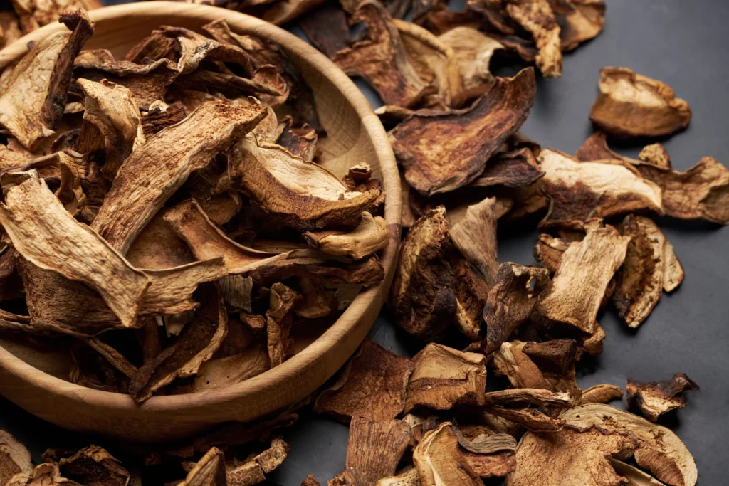 Fette di funghi essiccati in una ciotola di legno con altri pezzi sparsi su uno sfondo scuro